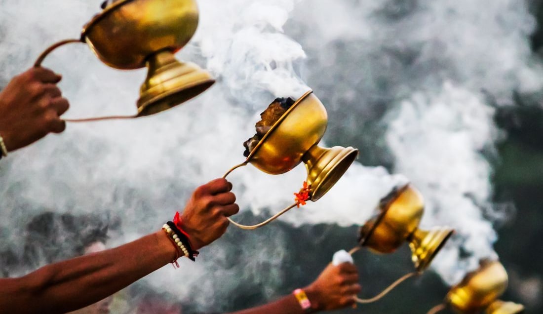  Close-up of multiple hands holding brass incense burners during a traditional Hindu aarti ceremony, with clouds of aromatic smoke rising in the air.