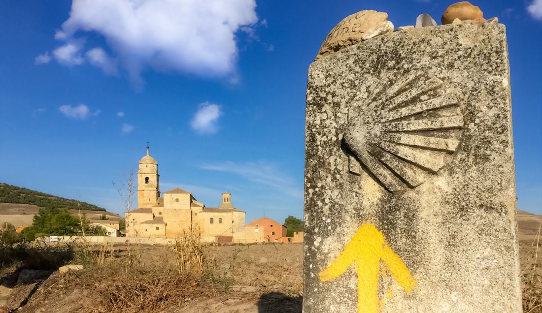 Stone waymarker with a carved scallop shell and yellow arrow pointing toward a church in Castrojeriz on the Way of St. James.